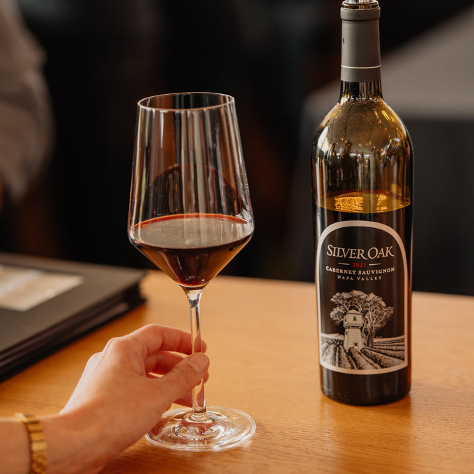 A bottle of Silver Oak Merlot wine with a pourer and a wine glass on a wooden table, with a hand holding the glass and a blurred restaurant background.