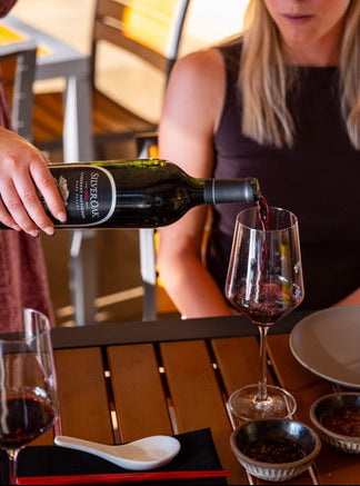 Person pouring red wine into a glass at an outdoor dining table.